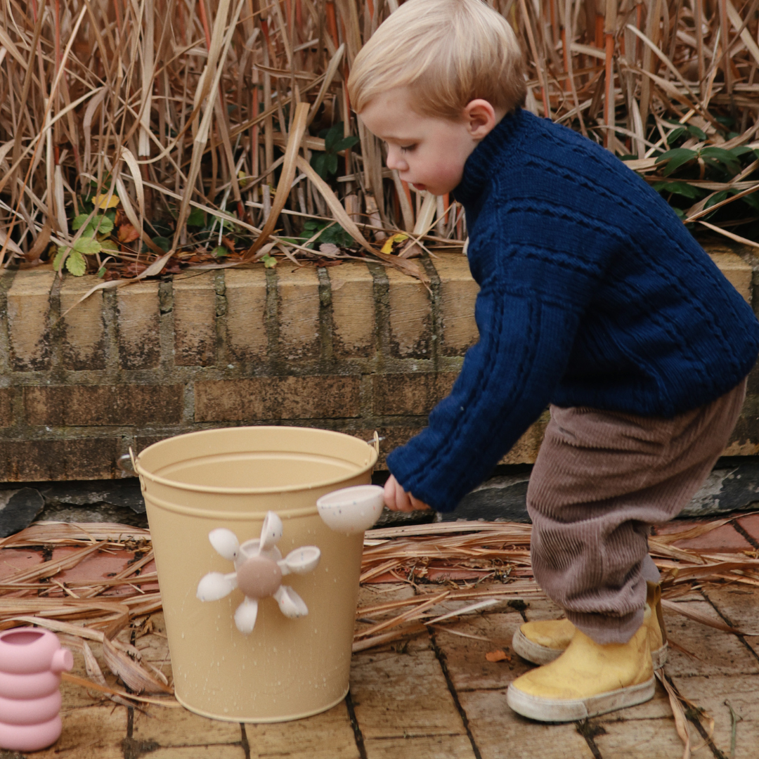Kisfiú sárga gumicsizmában a szabadban a vízkerék fürdőjátékkal játszik, amit egy vödör oldalára rögzített fel