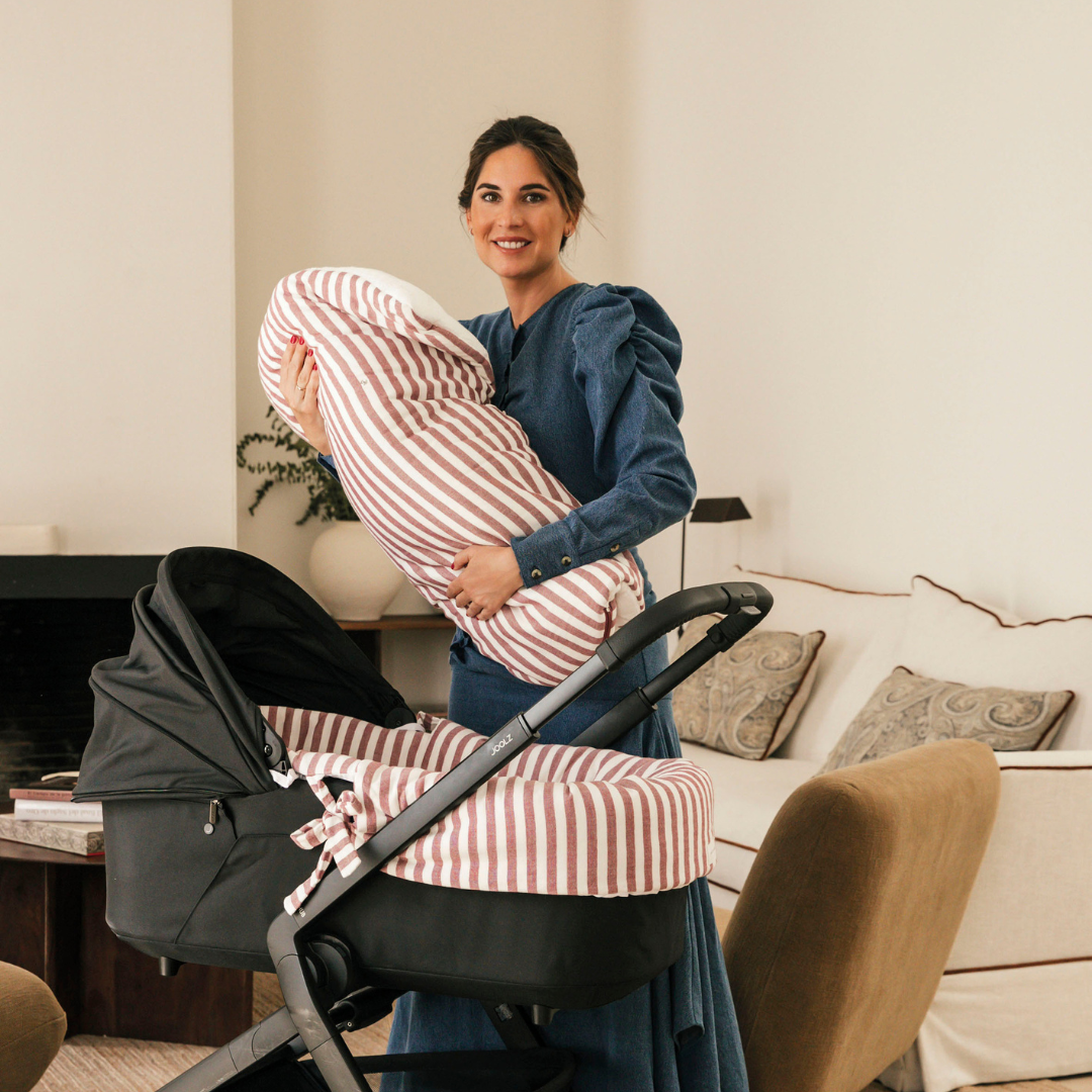 Édesanya a babakocsi mellett állva fogja kisbabáját a csíkos babafészek és takaróban