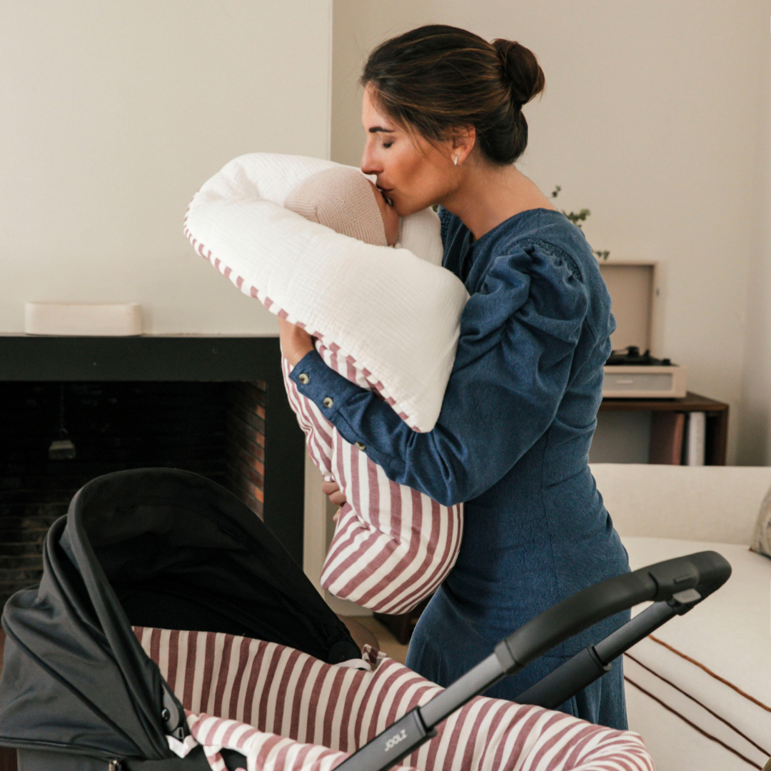 Édesanya megpuszilja a kisbabája homlokát, akit egy babafészek és takaróban tart magához közel. A babafészek és takaró mintája csíkos.