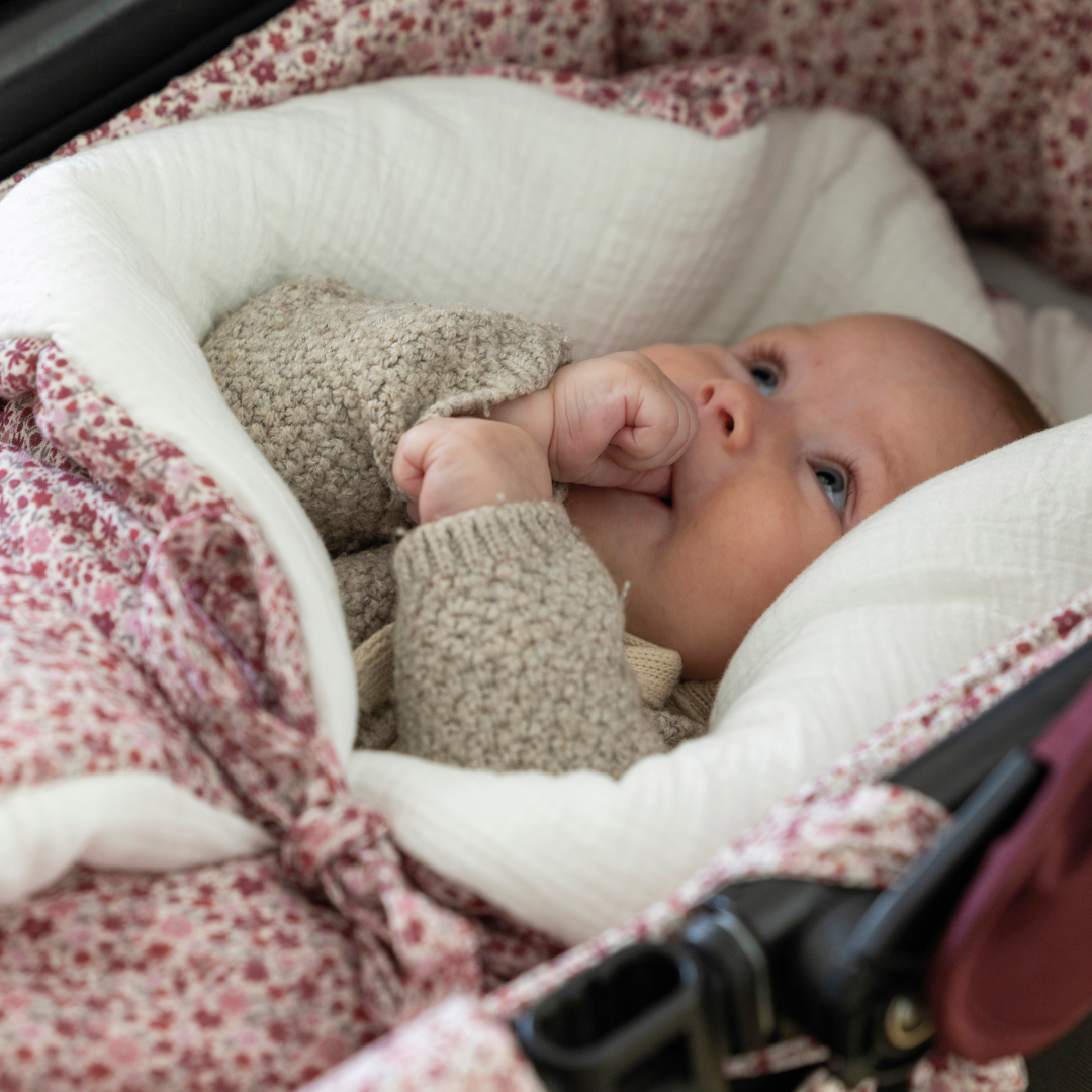 Kisbaba az ujját szopizza, miközben a virágmintás babafészekben pihen a babakocsiban