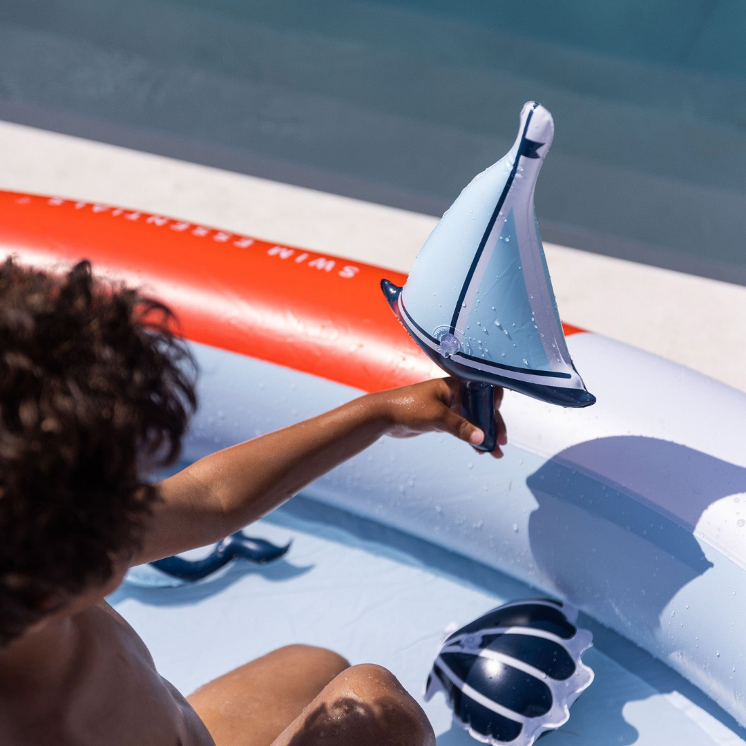 Gyermek játszik a medencéhez tartozó apró, kék-fehér felfújható vitorlás hajóval.