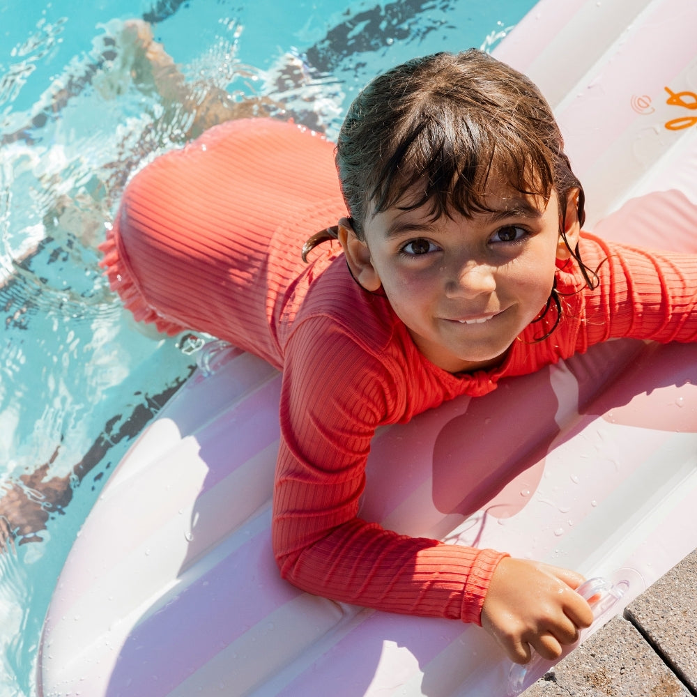 Kislány hasal a vízen a rózsaszín-fehér csíkos virágos felfújható szörfdeszkán.