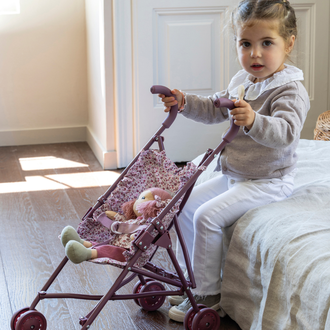 Kislány virágmintás játék babakocsival játszik a szobájában. A babakocsiban egy plüss baba ül.