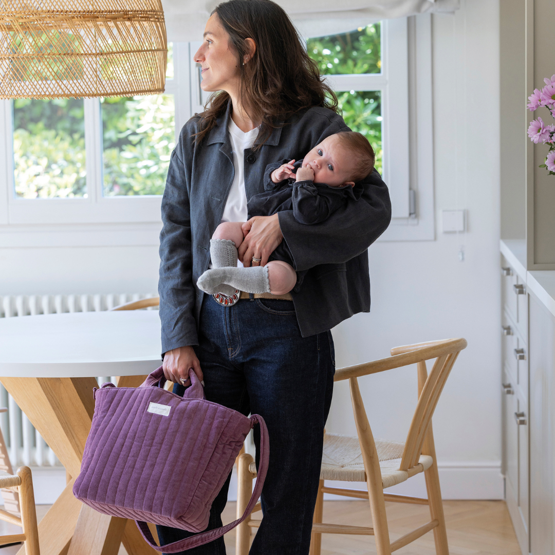 Édesanya kezében kisbabával bársony pelenkázó táskáját fogja a kezében, a konyhában állva
