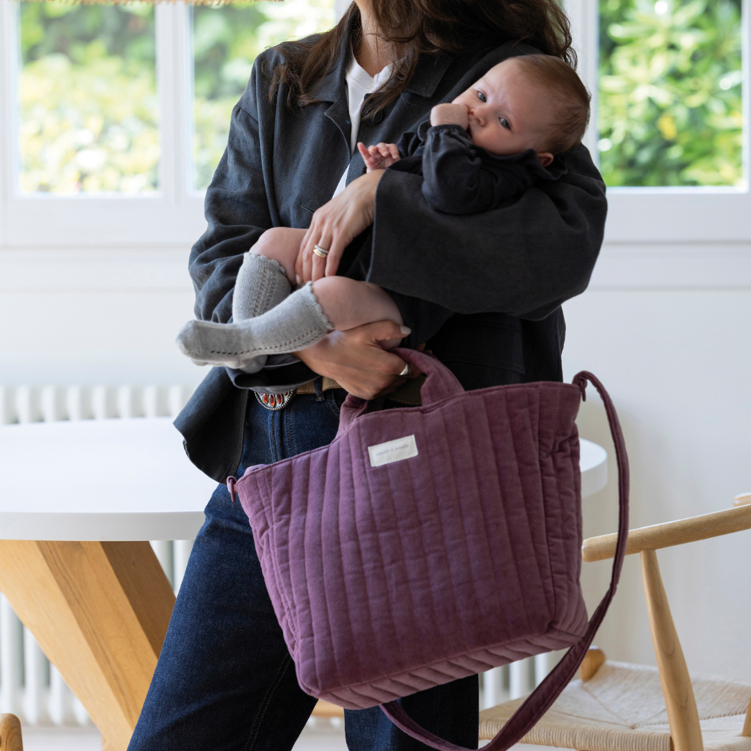 Édesanya kisbabát fog, miközben a kezében a rövid füleinél tartja a bársony pelenkázó táskáját