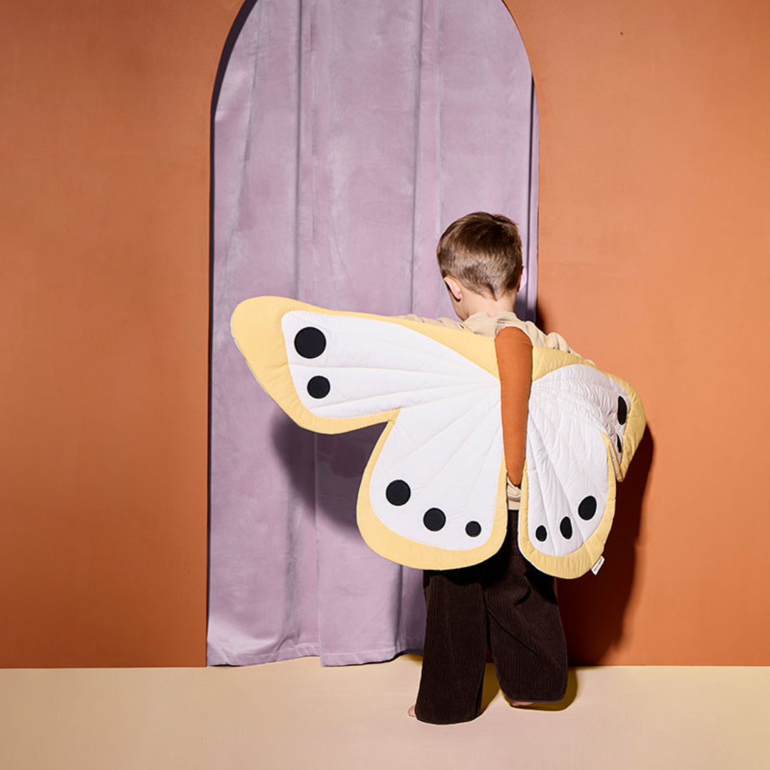 Kisgyermek sárga pillangó jelmezben, háttal állva lila függöny előtt