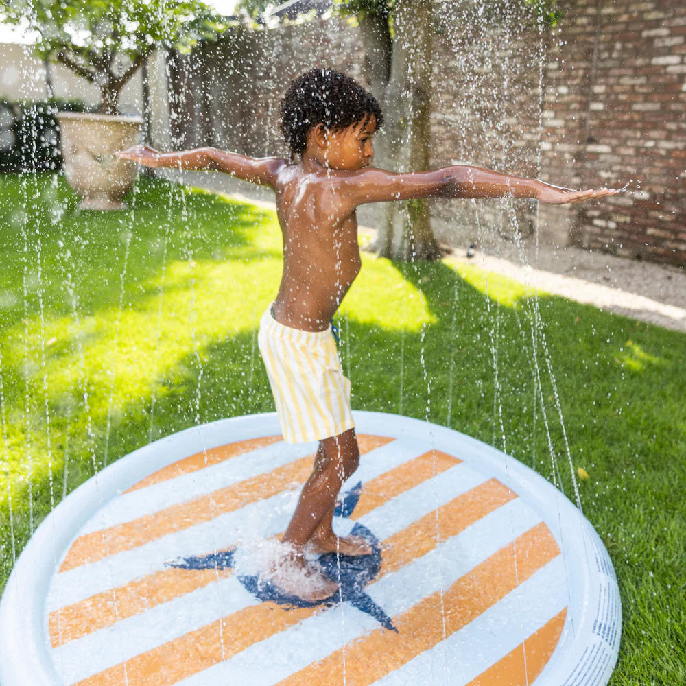 Kisfiú játszik kitárt karokkal a Swim Essentials trópusi cápás szökőkút pancsolójában a kertben.