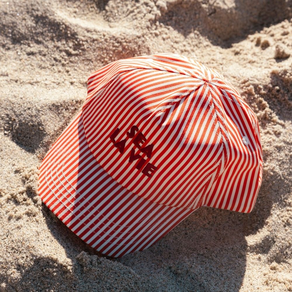 Swim Essentials csíkos gyerek baseball sapka a tengerparti homokban.