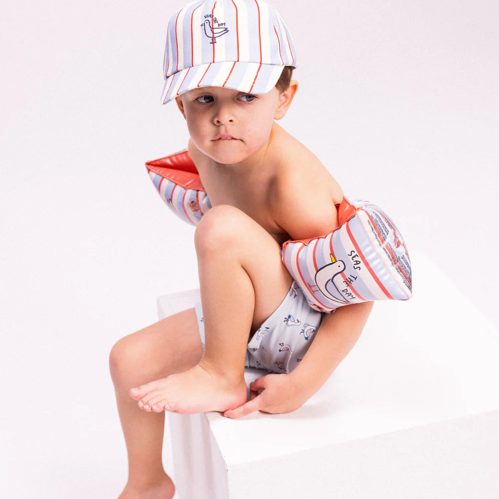 Kisfiú ül a stúdióban kék „Seas the Day” baseball sapkában és hozzá illő karúszóval.