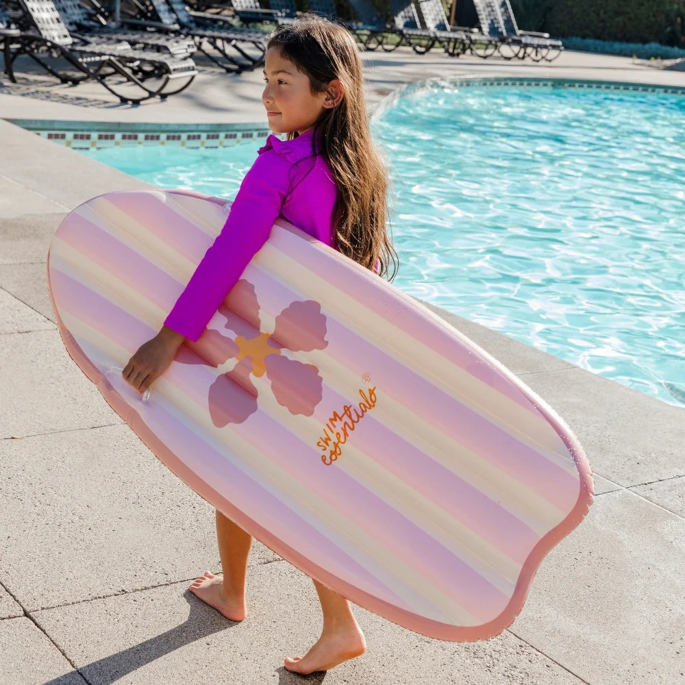 Kislány viszi a vállán a pasztell rózsaszín virágos Swim Essentials felfújható szörfdeszkát.