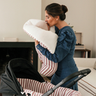 Édesanya megpuszilja a kisbabája homlokát, akit egy babafészek és takaróban tart magához közel. A babafészek és takaró mintája csíkos.