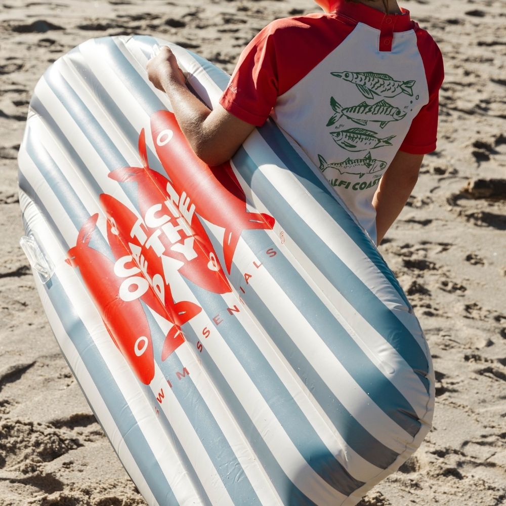 Gyermek viszi a vállán a piros halas mintás kék-fehér felfújható szörfdeszkát a homokos parton.