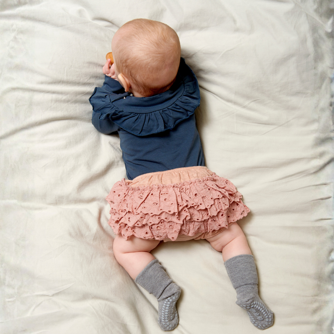 Kislány hason fekszik az ágyon rózsaszín szoknyában és szürke melange csúszásgátlós bambusz zokniban