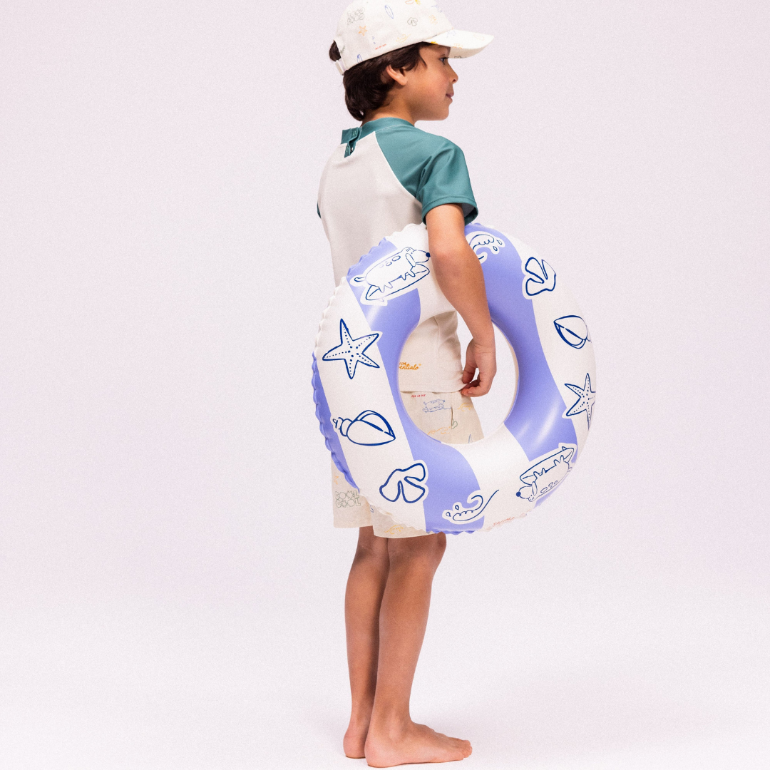 Kisfiú tengerparti szettben a kék csíkos szörfös úszógumival a karján.