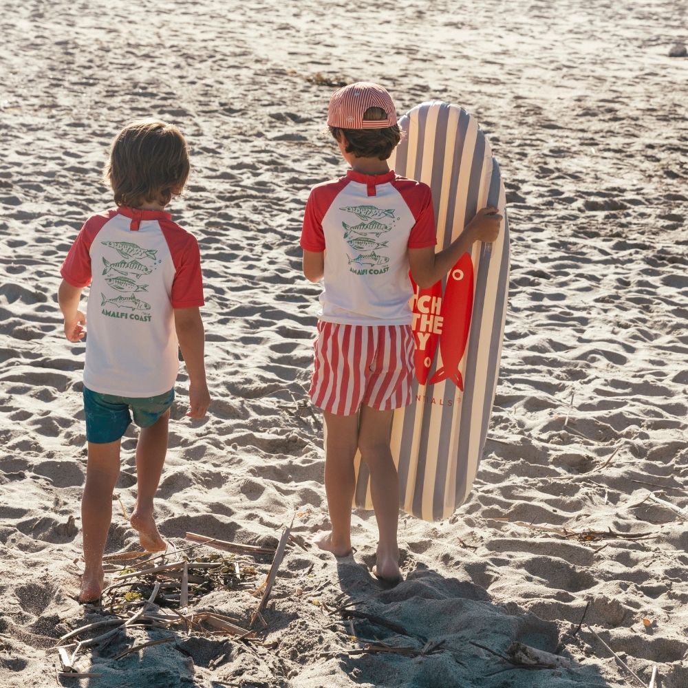 Két gyermek sétál a homokos tengerparton a kék-fehér csíkos Swim Essentials szörfdeszkával.