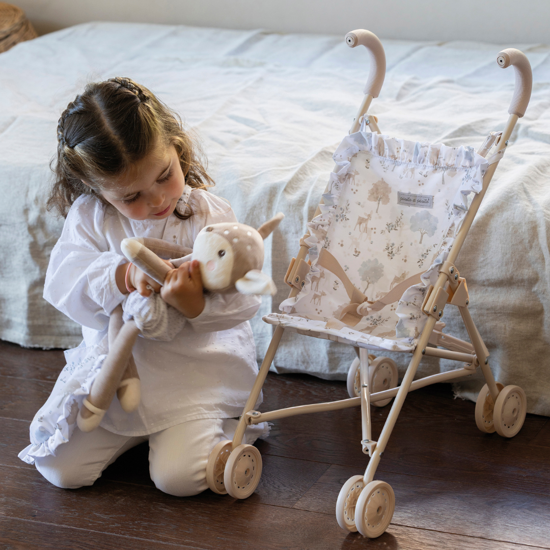 Kislány plüss őzikével játszik, mellette az összecsukható játék babakocsi Nara kollekciós mintákkal