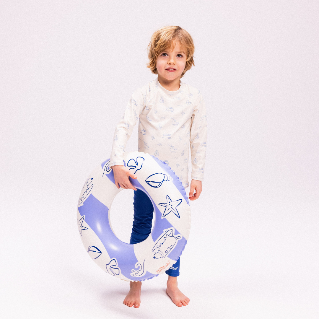 Kisfiú kezében a kék csíkos Swim Essentials gyerek úszógumival a tengerparti kalandokhoz.