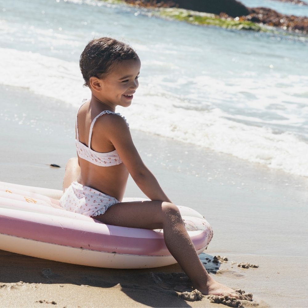 Kislány ül a tengerparti homokban a rózsaszín virágos Swim Essentials szörfdeszkán.