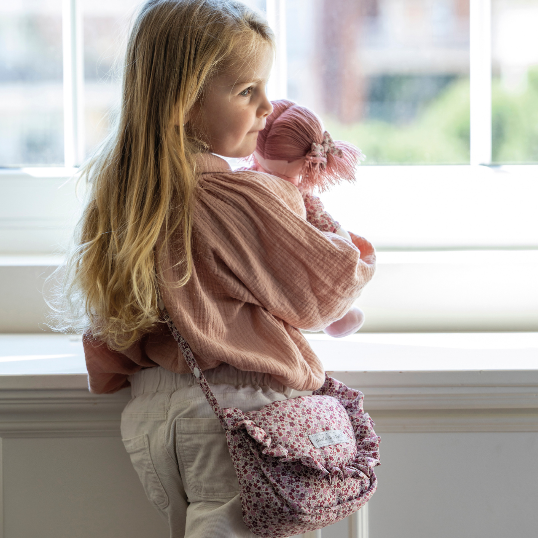 Kislány fodros válltáskával és plüss babával áll az ablak előtt - a fodros táska a klasszikus játék babakocsihoz tartozik