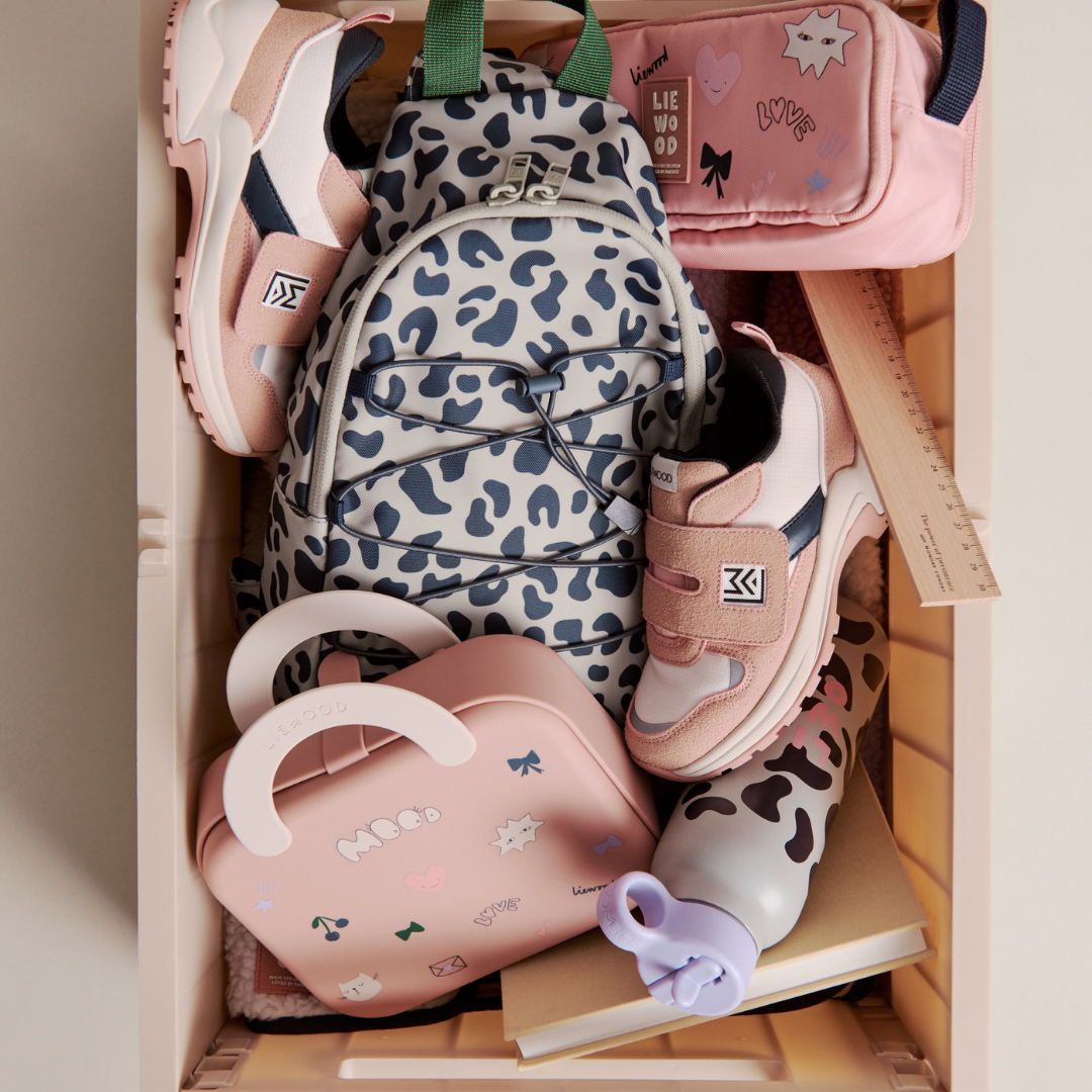 Egy tárolórekeszbe pakolt Liewood Falk gyerek kulacs pink kiegészítőkkel, mintás hátizsákkal és tornacipővel felülnézetből
