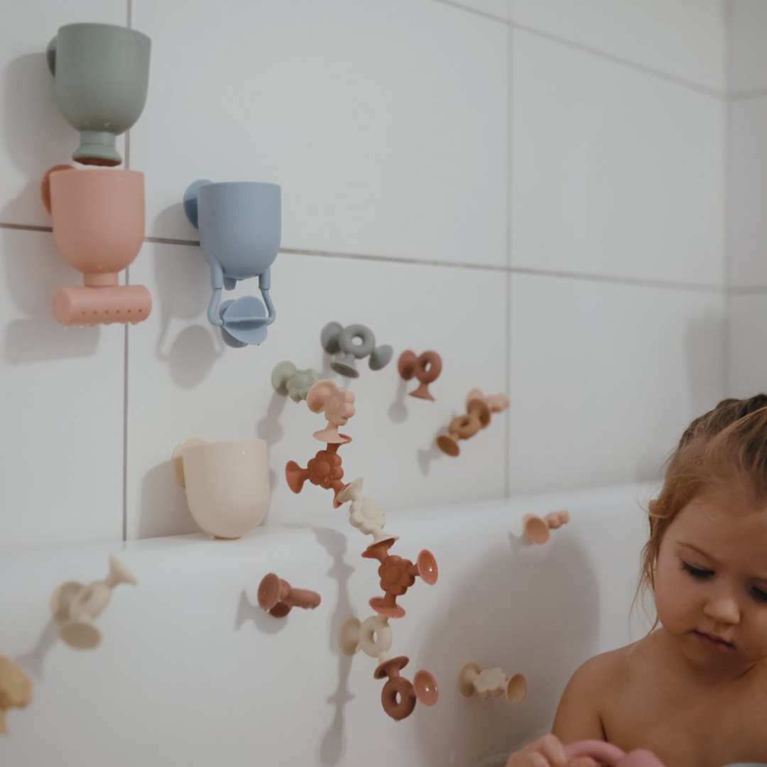 Mushie Önts és játssz! fürdőjáték tapadókorongokkal a fürdőszoba csempéjére rögzítve, háttérben fürdő gyermekkel