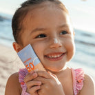 Kislány mutatja a Swim Essentials 50 ml-es, érzékeny bőrre kifejlesztett naptejét a strandon.