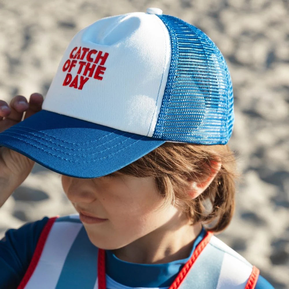 Kisfiú tengerparti homokban kék-fehér hímzett "Catch of the Day" Swim Essentials baseball sapkában, UPF 50+ védelemmel.