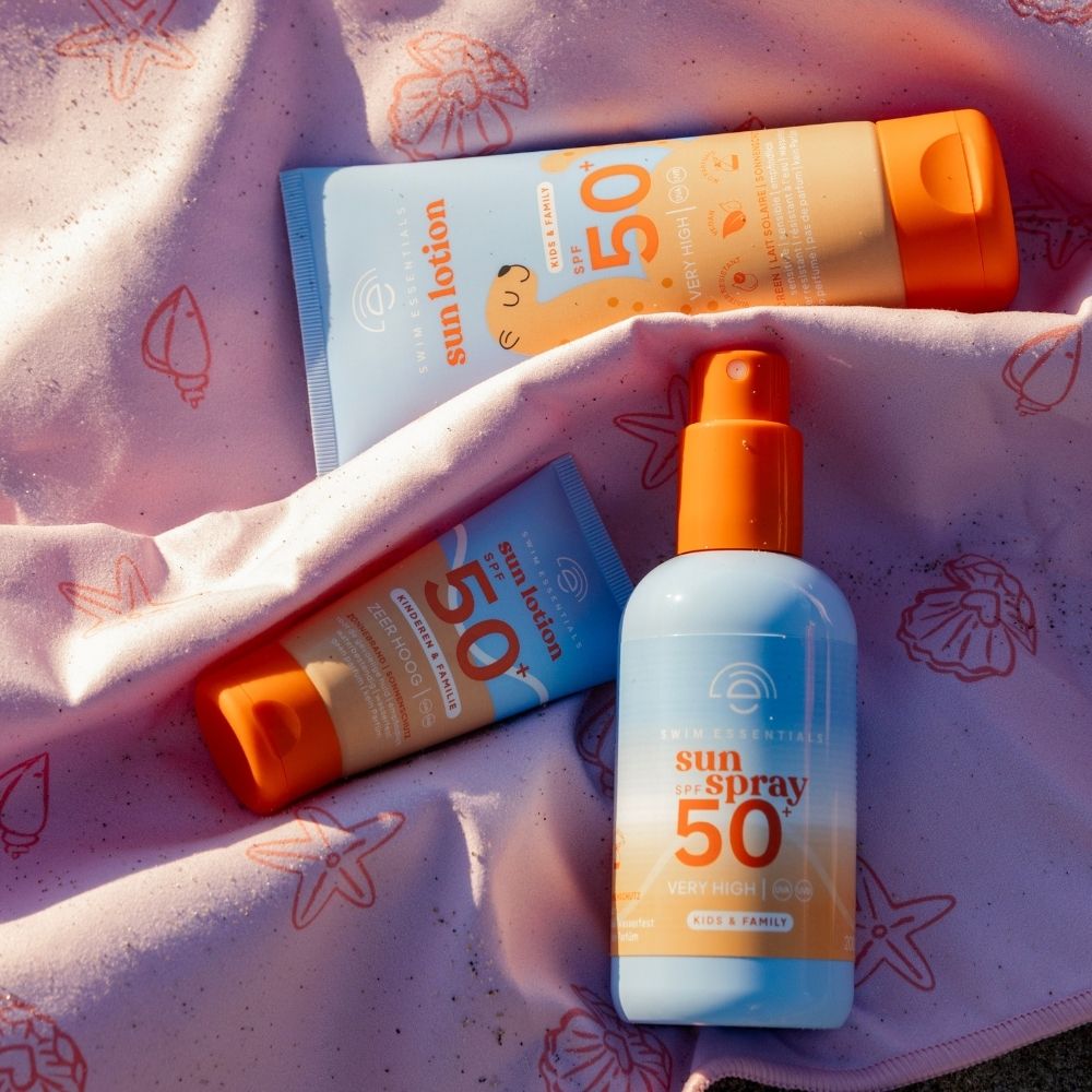 Swim Essentials napvédő termékek: spray és tubusos naptejek egy tengerparti törölközőn.