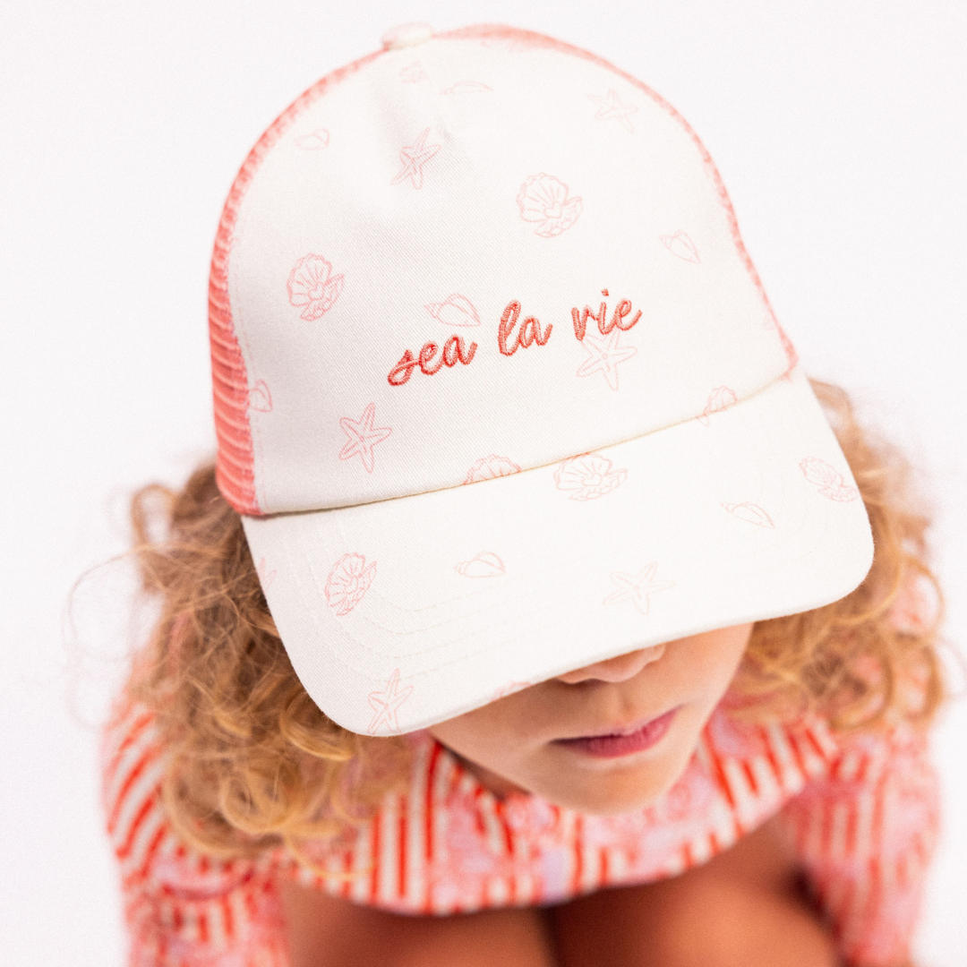 Kislány felülről kagyló mintás Sea La Vie baseball sapkában és piros csíkos úszóruhában.