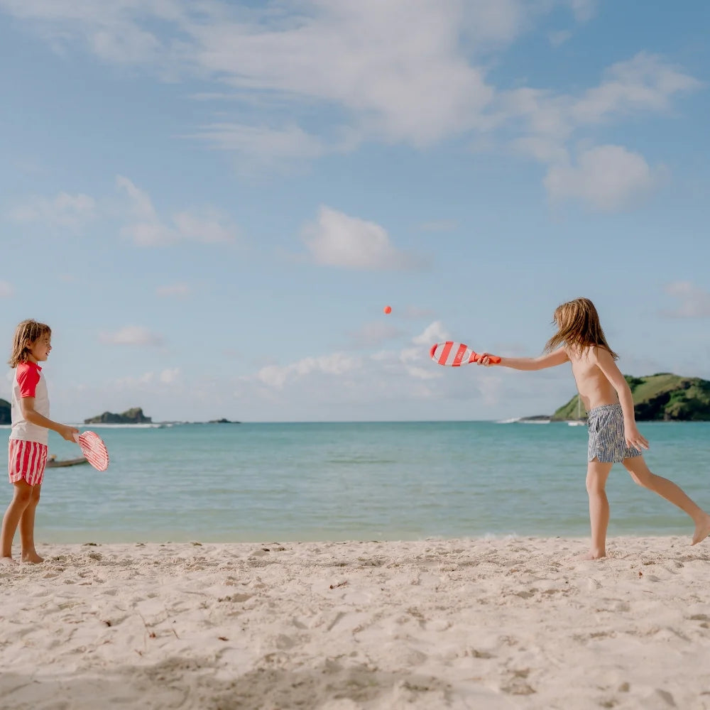 Két kisgyerek önfeledten játszik a Swim Essentials homár mintás strandtenisz szettel a homokos tengerparton, kék égbolt és tenger előtt