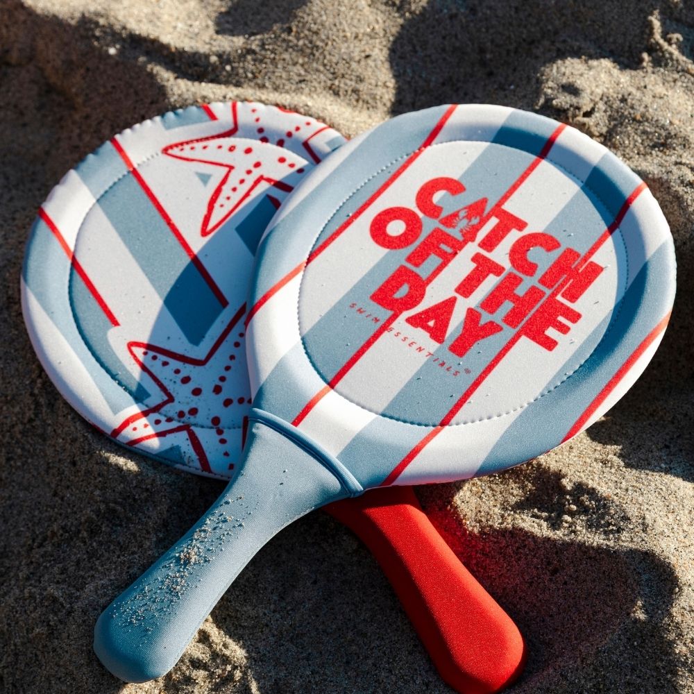 A két Swim Essentials strandtenisz ütő stílusosan a homokba fektetve a tengerparton