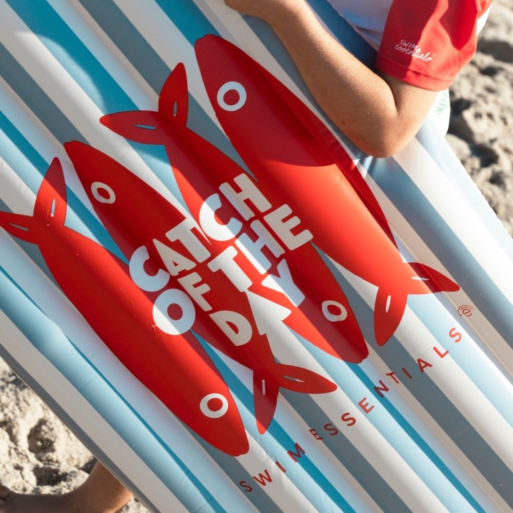 Közelkép a "Catch of the Day" feliratú piros halas mintáról a kék csíkos szörfdeszkán.
