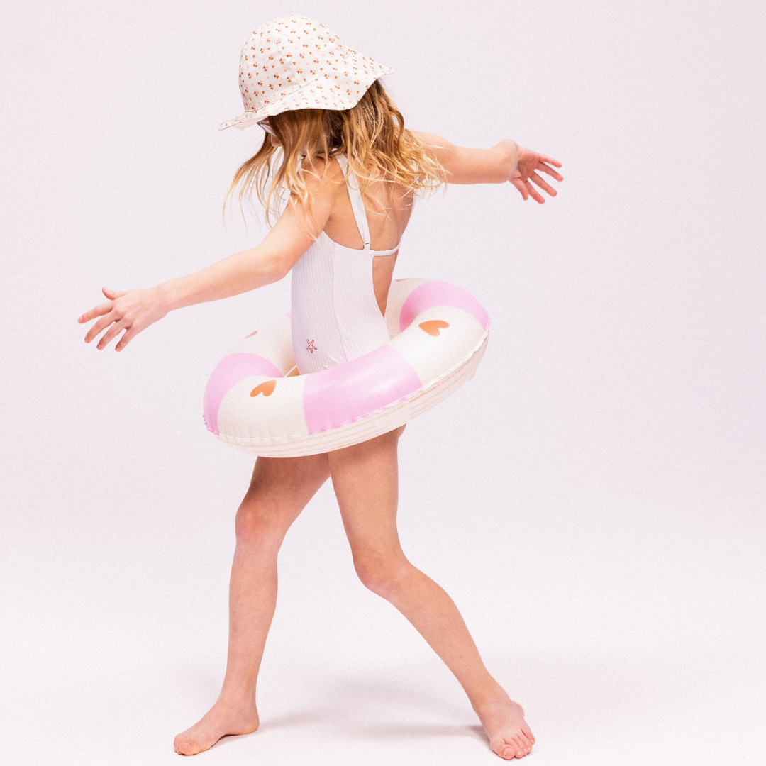 Kislány strandkalapban és fehér fürdőruhában pózol a rózsaszín szívecskés Swim Essentials úszógumival.