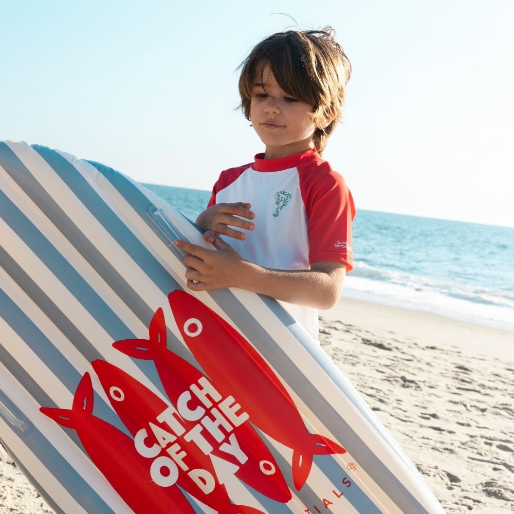 Gyermek áll a tengerparton a 120 cm-es kék-fehér csíkos felfújható szörfdeszkával.