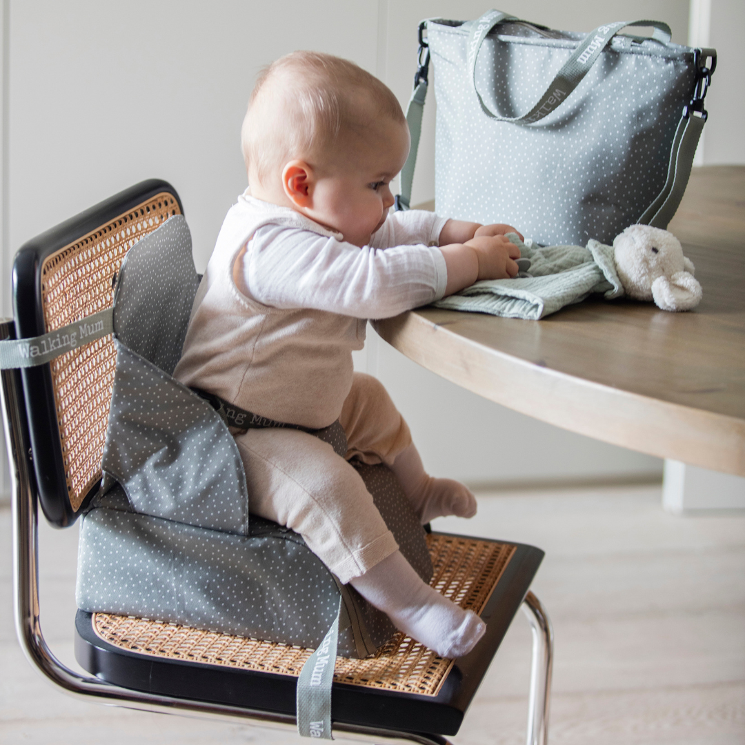 Pippadu oliva zöld pottyös textil székmagasító székre rögzítve, benne ülő babával és a hozzá tartozó hordozótáskával az asztalon